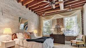 Un dormitorio con pared de ladrillo, techo de madera, cama con ropa de cama blanca, dos mesitas de noche con lámparas, cómoda, una silla y ventana con persianas.