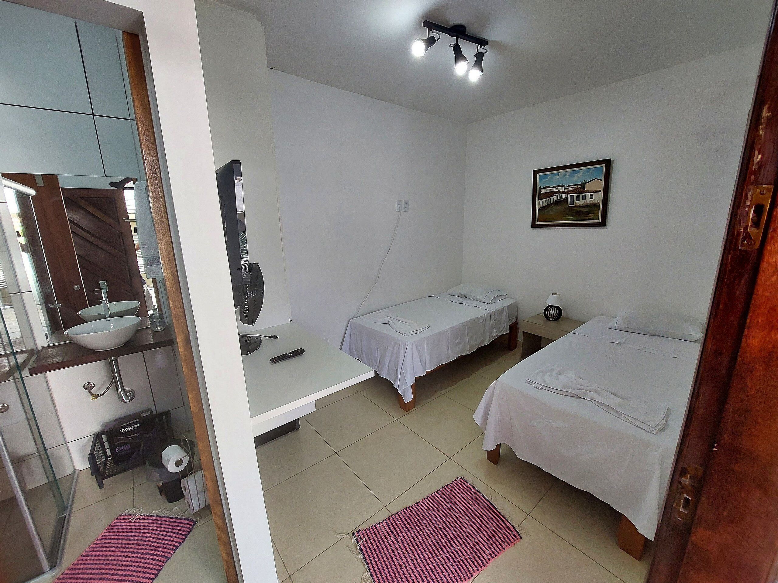 A small, clean room with two single beds, a bedside table with a lamp, a small desk with a computer and fan, a bathroom with a sink and toilet, and a picture on the wall.