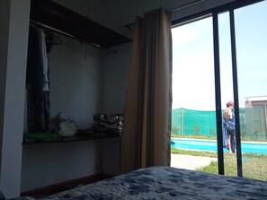 A room with a bed, a shelf with clothes, and a view of a swimming pool.