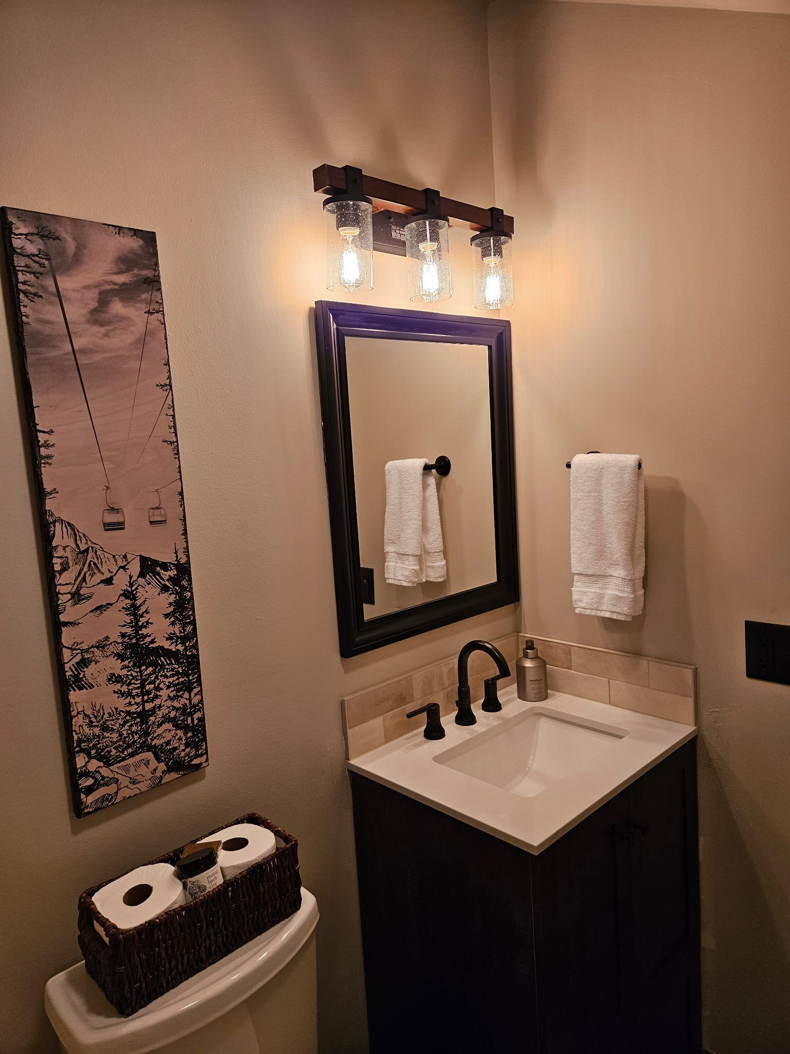 Une salle de bain avec un lavabo, un miroir et un porte-serviettes. Il y a un tableau accroché au mur et un panier de papier toilette sur les toilettes.
