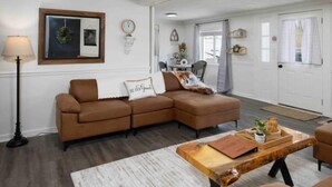 Ein Wohnzimmer mit einem braunen Ecksofa, einem Couchtisch und einer Lampe. Im Hintergrund ist ein Essbereich mit Tisch und Stühlen zu sehen.