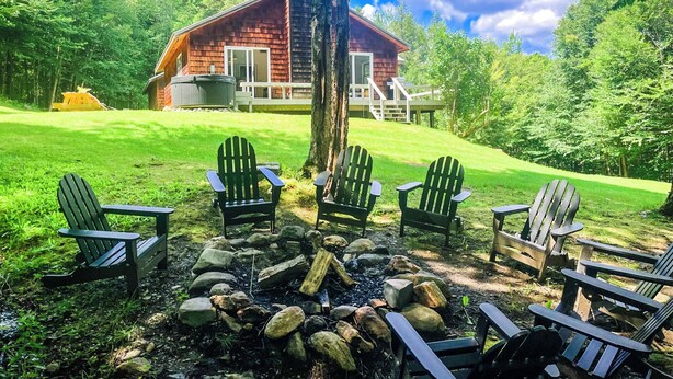 En stuga med en veranda, omgiven av en gräsyta med Adirondack-stolar och en eldstad.