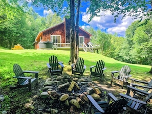 Eine Holzhütte mit einer Veranda, umgeben von einer Wiese mit Adirondack-Stühlen und einem Feuerkorb.