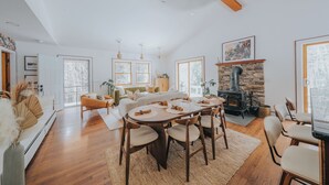 Ett rymligt vardagsrum med ett stort matbord i trä, bekväma sittplatser, en eldstad i sten och stora fönster som släpper in naturligt ljus.