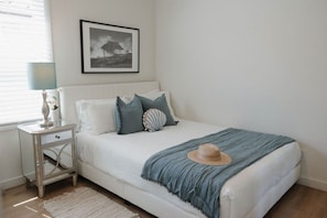 A neatly arranged bedroom with a bed, pillows, a bedside table with a lamp, a framed picture on the wall, and a window with blinds.