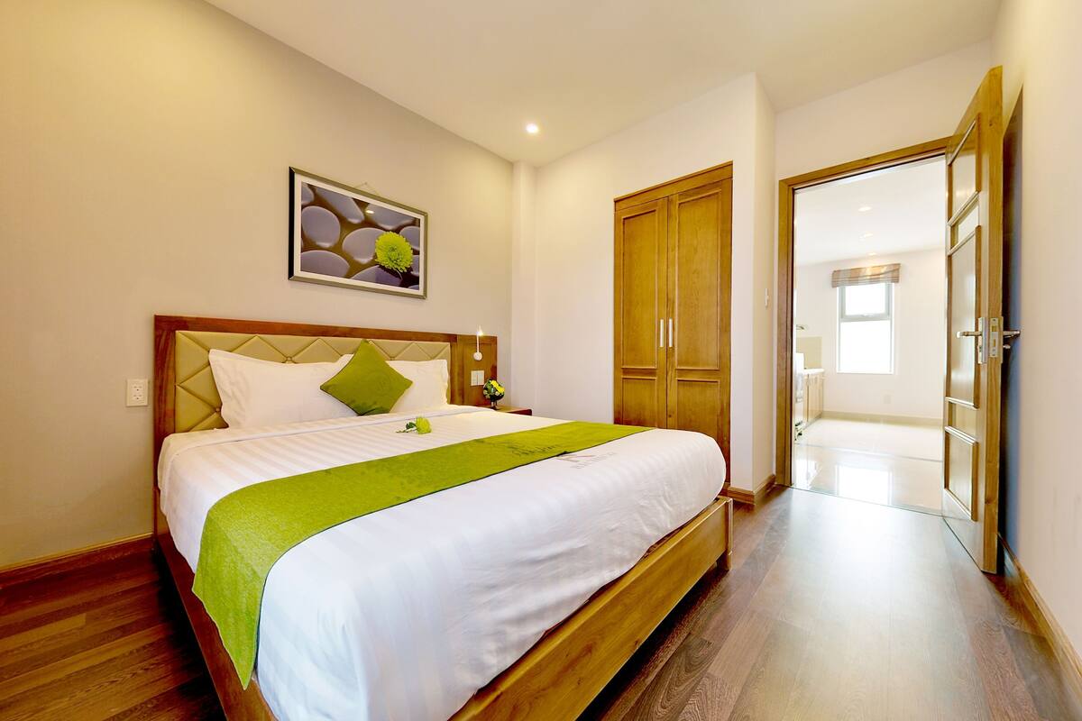 Une chambre d’hôtel avec un lit, un plaid vert, un tableau encadré, une armoire en bois et un couloir menant à une autre pièce.