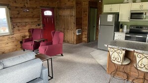 A log cabin interior with a living area featuring a sofa and armchairs, a kitchen with a refrigerator, microwave, and stove, and a bar area with stools. - Camas Creek Cottage (Hamilton)