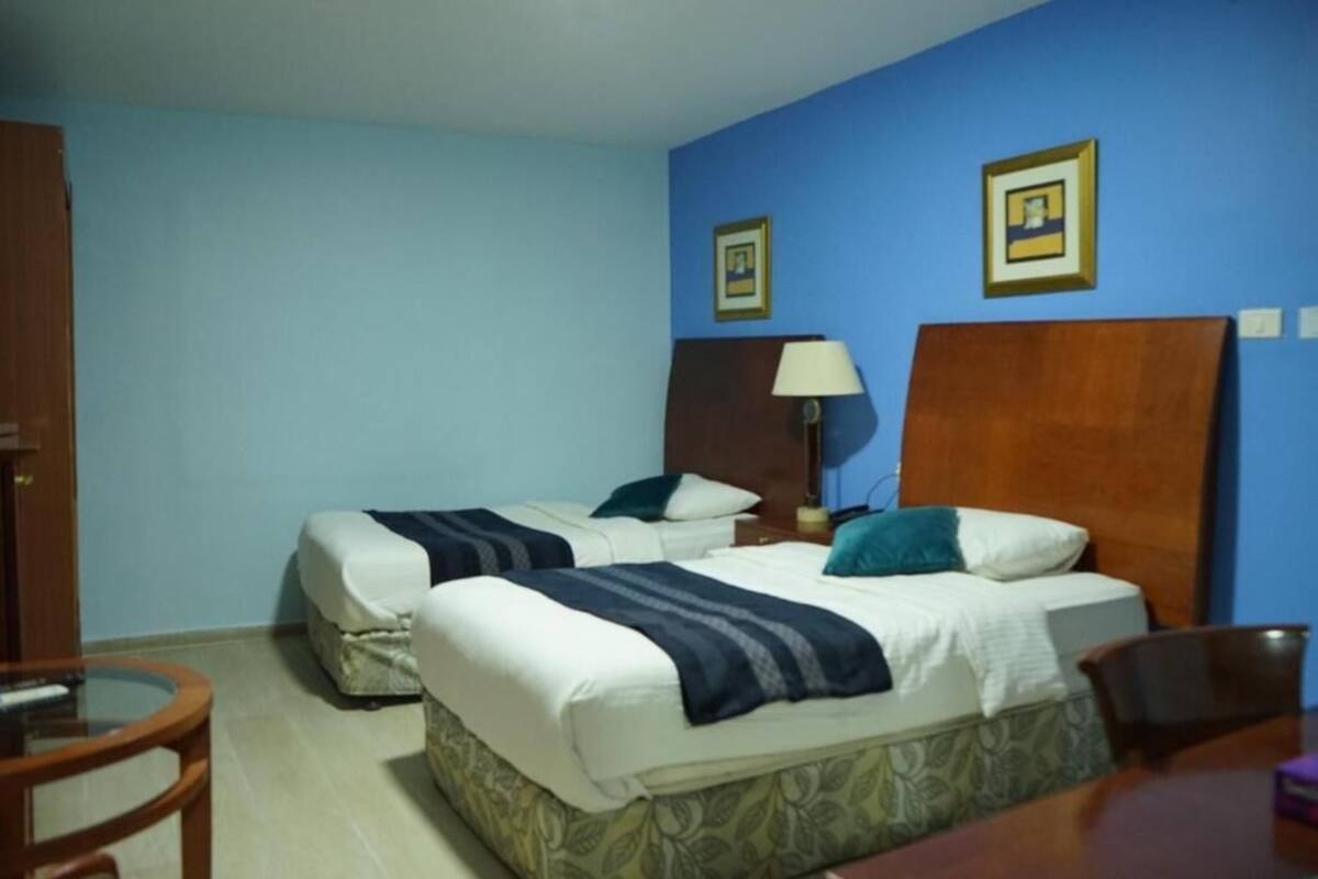 A room with two beds, a wooden headboard, a nightstand with a lamp, and a small round table.
