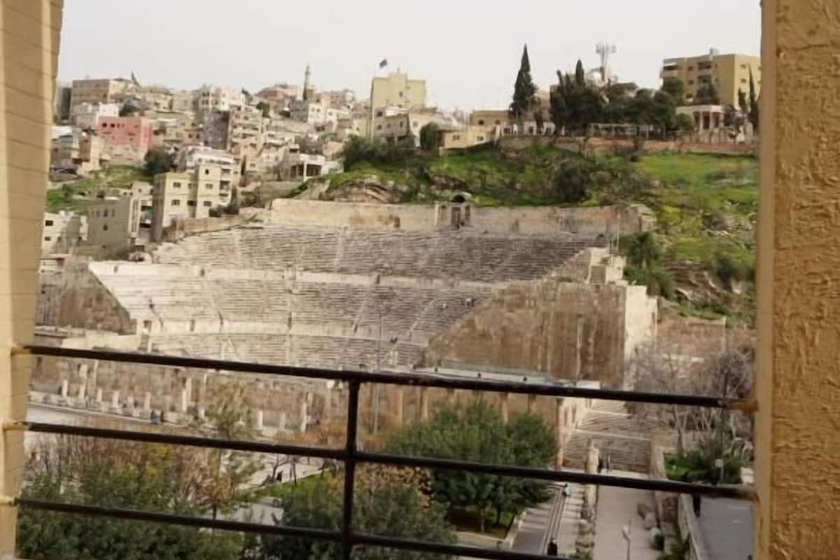 A view from a window with a metal railing, overlooking a densely built urban area with various buildings and some greenery.
