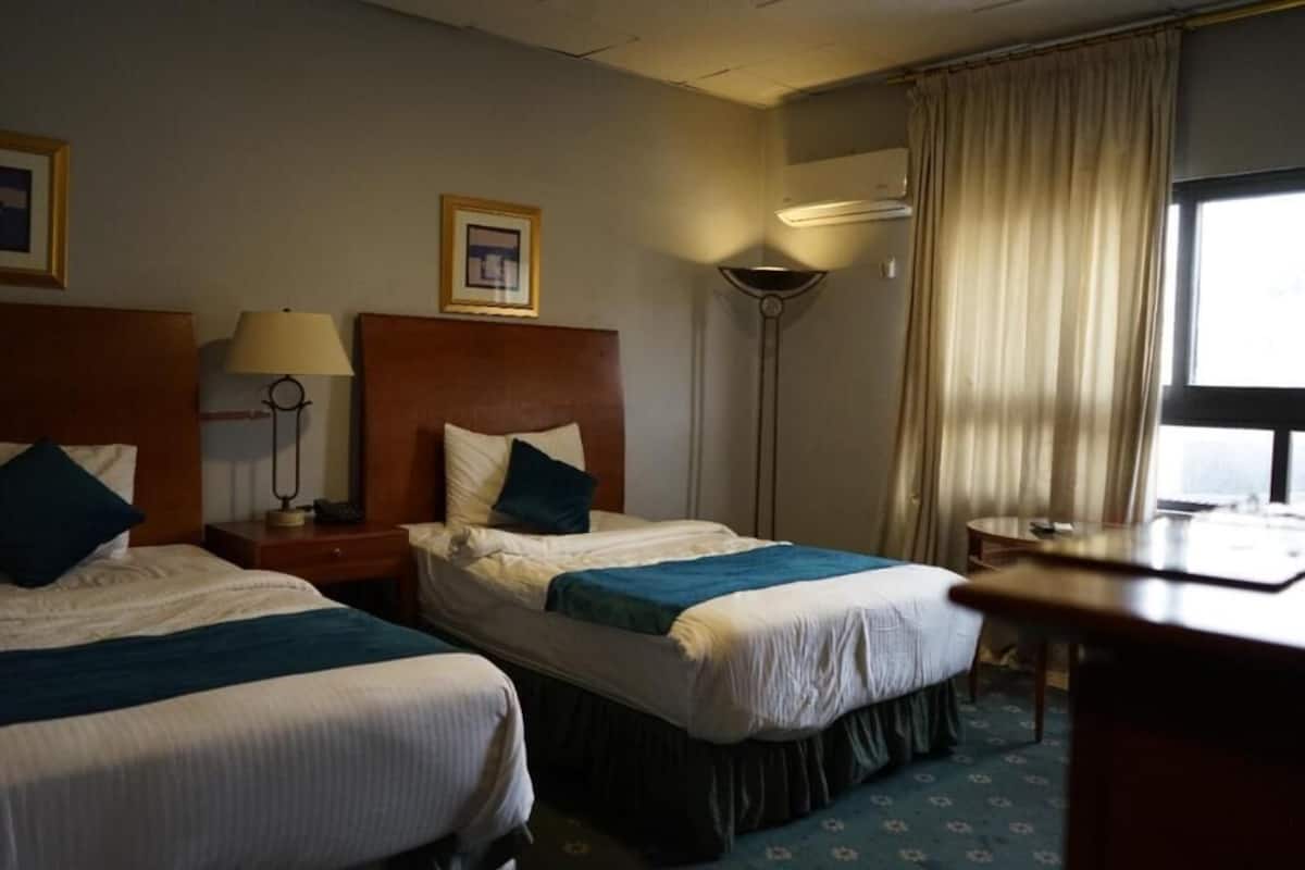 A hotel room with two beds, a desk, a lamp, a picture frame, and a window with curtains.