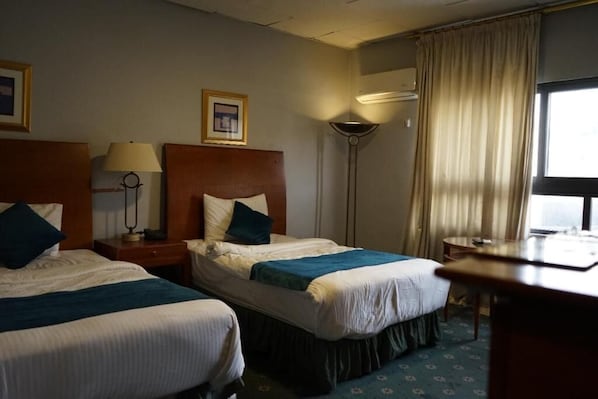 A hotel room with two beds, a desk, a lamp, a picture frame, and a window with curtains.