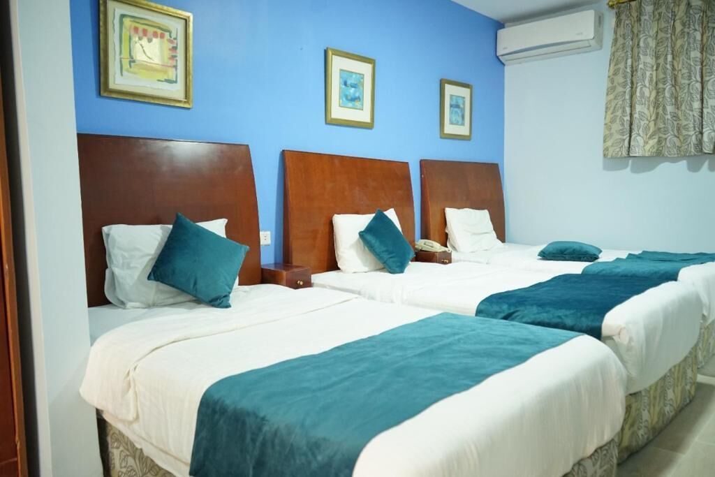 A hotel room with two beds, each with a wooden headboard, white bedding, and teal throw blankets. There are framed pictures on the wall, a bedside table with a lamp, and an air conditioning unit on the wall.