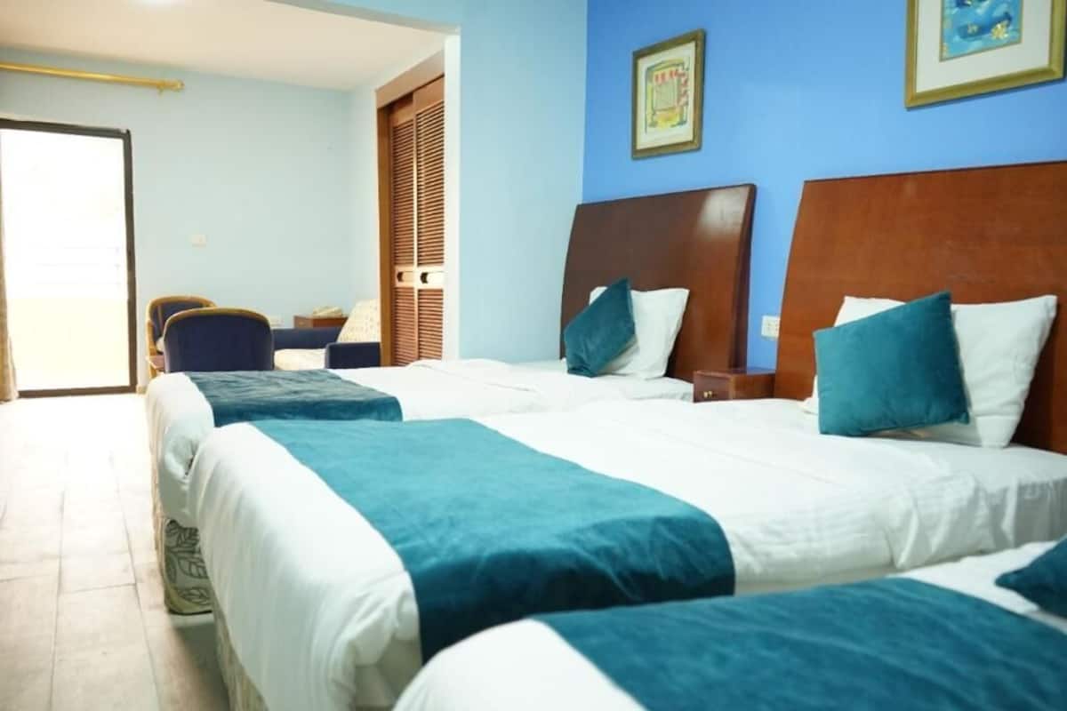 A hotel room with two beds, each with a wooden headboard, white bedding, and teal throw pillows. There is a small table with a chair, a window with blinds, and a door leading outside.