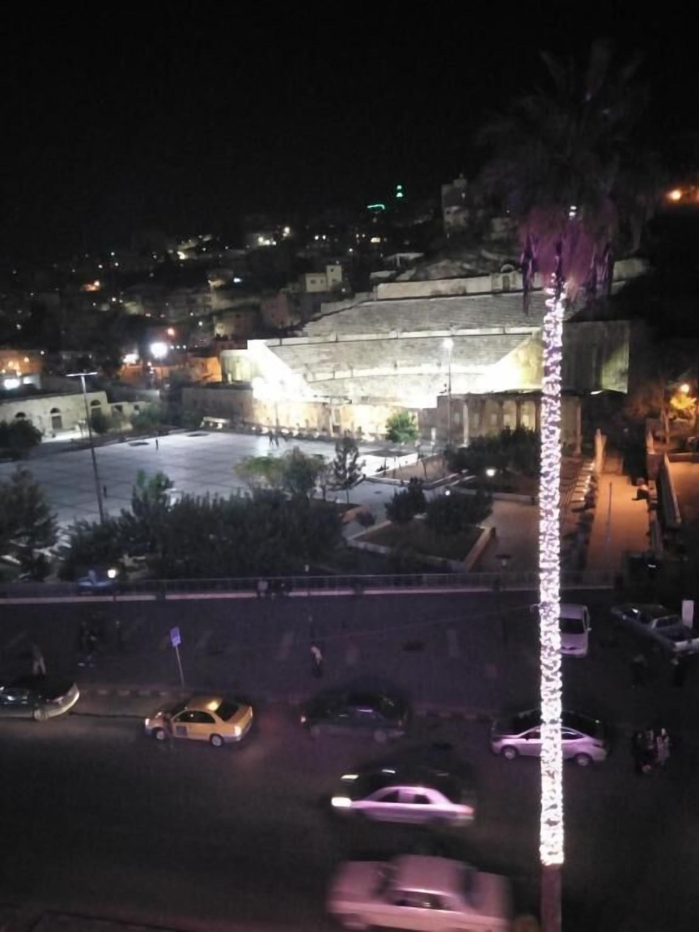 A nighttime view of a cityscape with a prominent building featuring a large open plaza and a tall, illuminated structure.