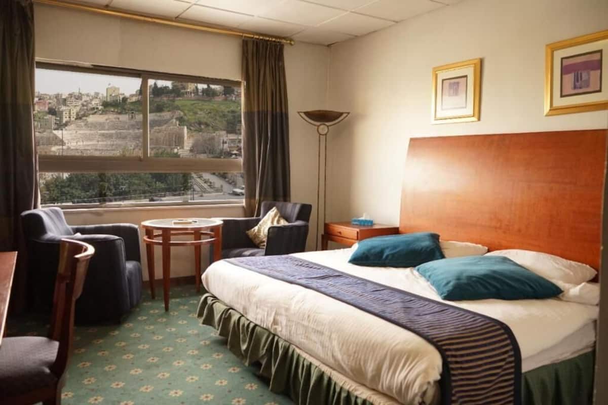 A hotel room with a large bed, a chair, a small table, a lamp, and a view of a cityscape through the window.