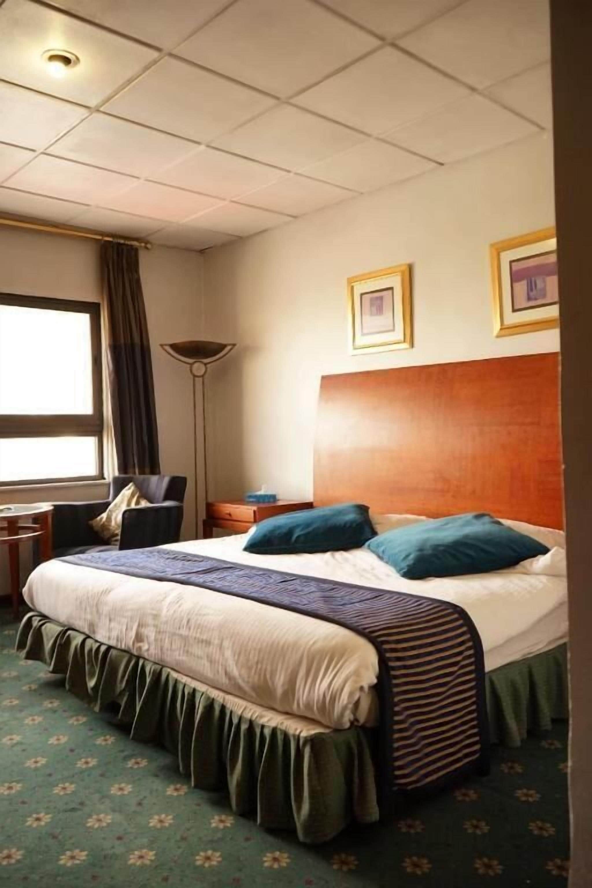 Una camera d'albergo con un letto grande, una scrivania, una sedia, una lampada da terra e una finestra con tende.