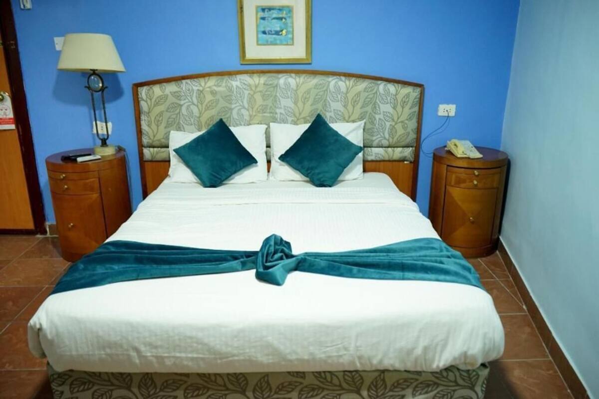A neatly made bed with a teal bedspread and pillows, a bedside lamp, and a framed picture on the wall.