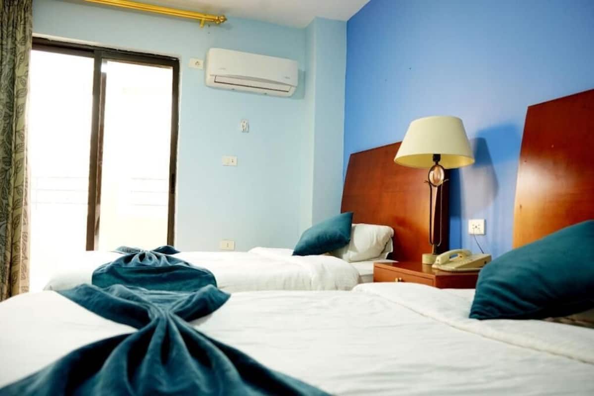 A hotel room with two beds, a bedside table with a lamp, an air conditioner, and a window with curtains.