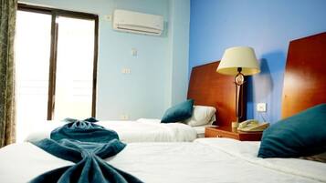A hotel room with two beds, a bedside table with a lamp, an air conditioner, and a window with curtains.