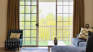 A room with a sliding glass door leading to a balcony with a white railing. There is a chair with a patterned cushion and a sofa with a cushion. A small table is on the balcony.