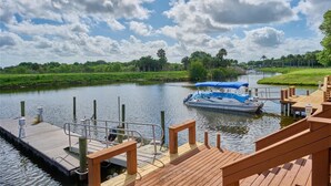 A wooden dock with a boat, a calm river, and a grassy landscape with trees.