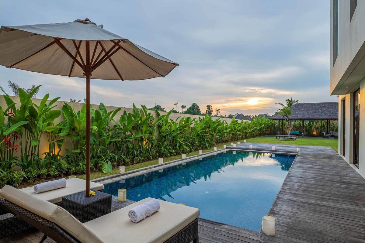 Une piscine entourée de chaises longues et d’un parasol, le tout bordé d’une végétation luxuriante et d’une terrasse en bois.