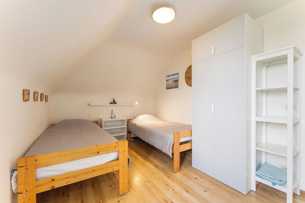 Un dormitorio pequeño y bien iluminado con dos camas de madera, un armario blanco y una estantería con una lámpara.