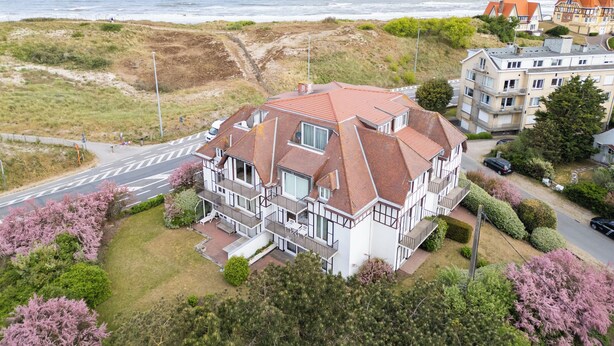 A coastal residential building with a red-tiled roof, white walls, and multiple balconies. It is situated near a beach with a road in the foreground.