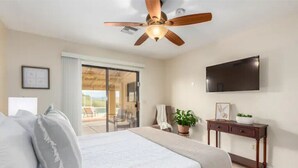 A bedroom with a bed, a ceiling fan, a TV mounted on the wall, a sliding glass door leading to a balcony, and a small table with a plant.