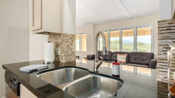 A modern kitchen with a double sink, a stone backsplash, and a view of a couch and outdoor scenery through large windows.