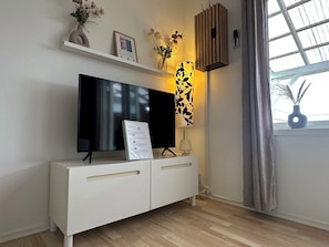 A modern living room with a flat-screen TV on a white cabinet, a decorative wall piece, and a window with curtains.
