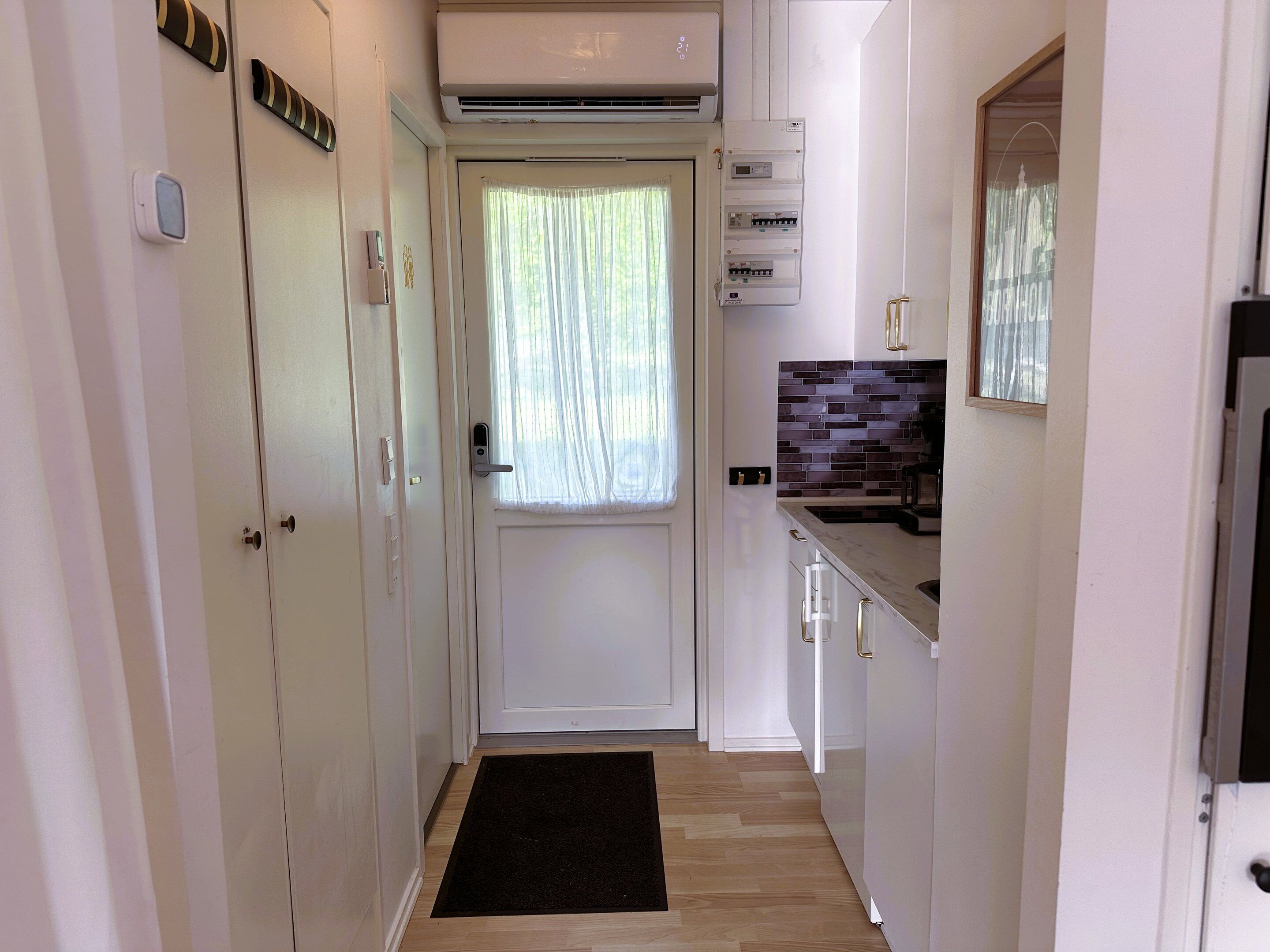 A narrow hallway with a door, a black doormat, a tiled backsplash, and a small kitchen area with cabinets and a countertop.