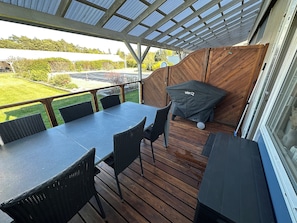 A covered outdoor patio with a table and chairs, a grill, and a view of the surrounding area.