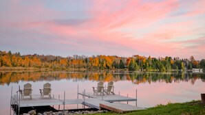 A serene lakeside scene with two Adirondack chairs on a wooden dock, surrounded by autumn-colored trees.