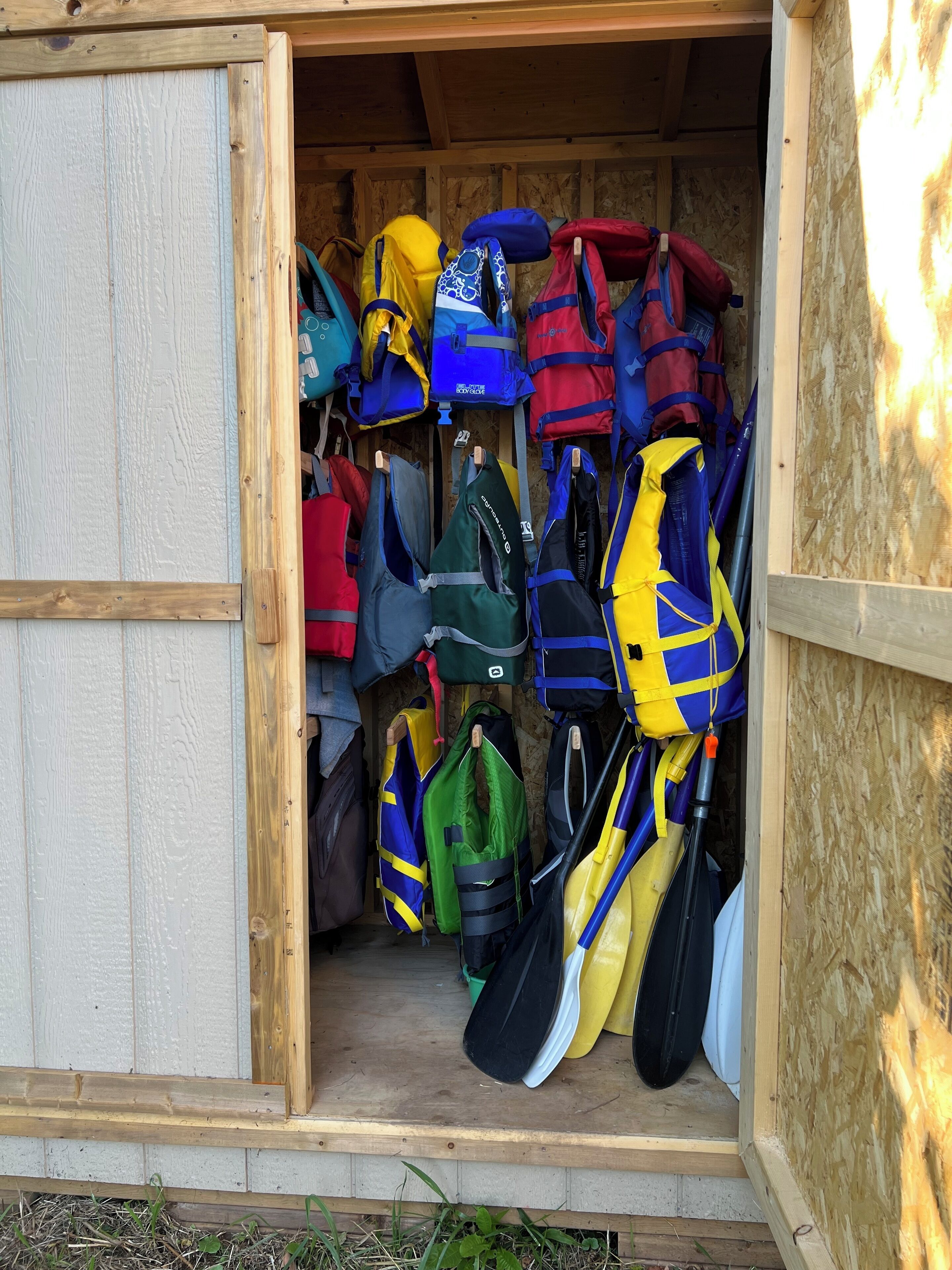 A wooden structure with a door open, revealing a collection of colorful life jackets and paddles hanging inside.