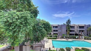 A swimming pool surrounded by a building with multiple balconies and a large tree in the foreground. - Charming & Comfy Condo in the Heart of Old Town! (Scottsdale)