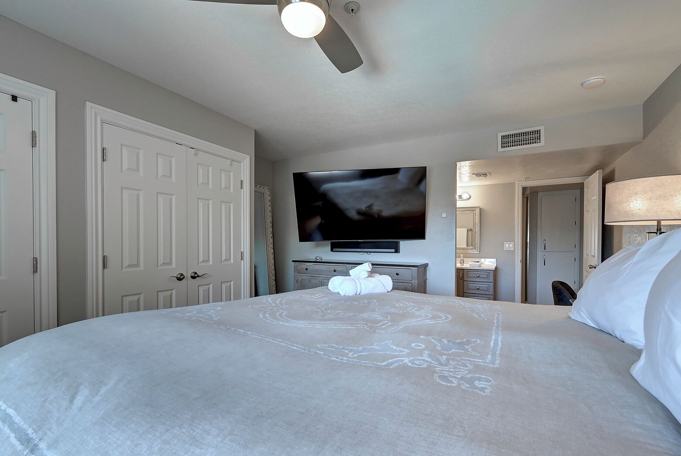 A hotel room with a large bed, a ceiling fan, a flat-screen TV mounted on the wall, and a bathroom visible through an open door.