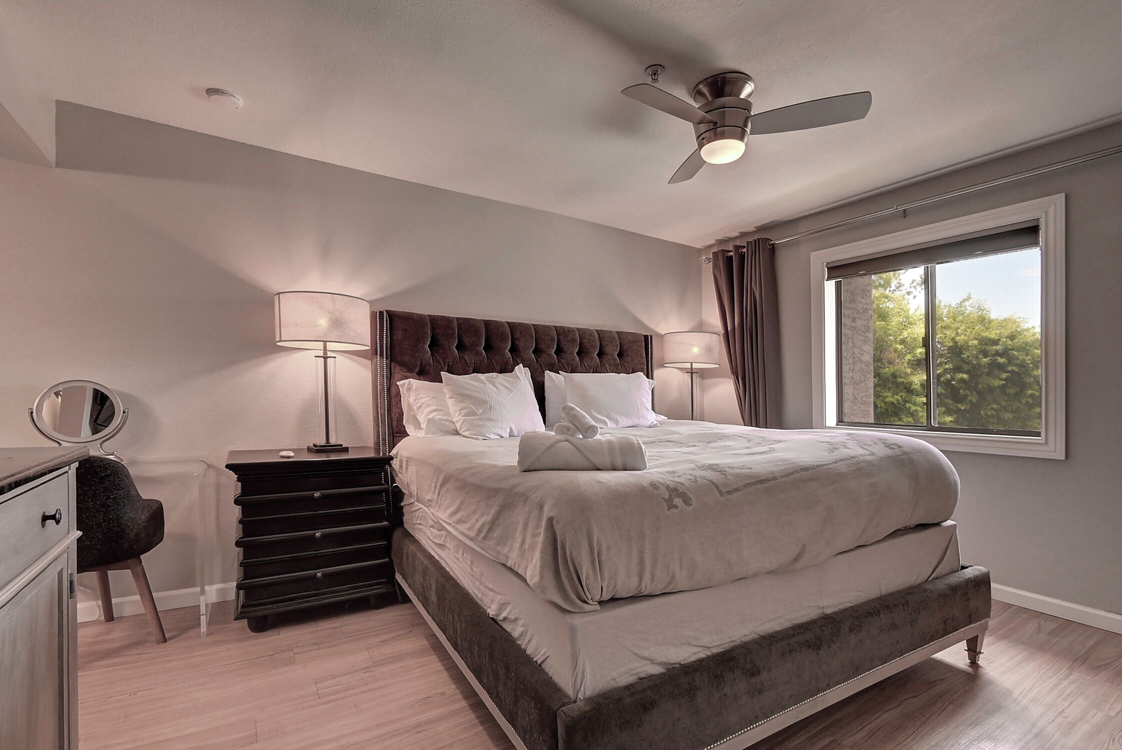 A bedroom with a large bed, a ceiling fan, a dresser, a vanity with a mirror, and a window with a view of trees.
