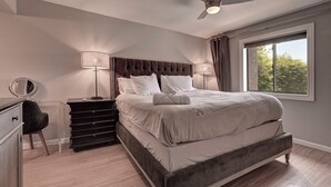 Un dormitorio con una cama grande, un ventilador de techo, un aparador, una tocador con espejo y una ventana con vistas a árboles.