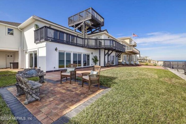 A two-story house with a balcony, a patio with outdoor furniture, and a view of the ocean.