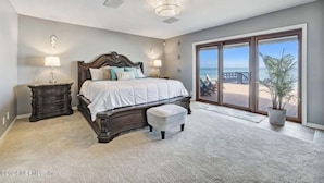 Ein Schlafzimmer mit einem großen Bett, zwei Nachttischen, einem Fenster mit Blick auf den Strand und einer Glastür, die zu einem Außenbereich führt.