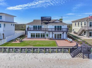A two-story house with a balcony, a patio with outdoor furniture, and a sandy beach in the background.