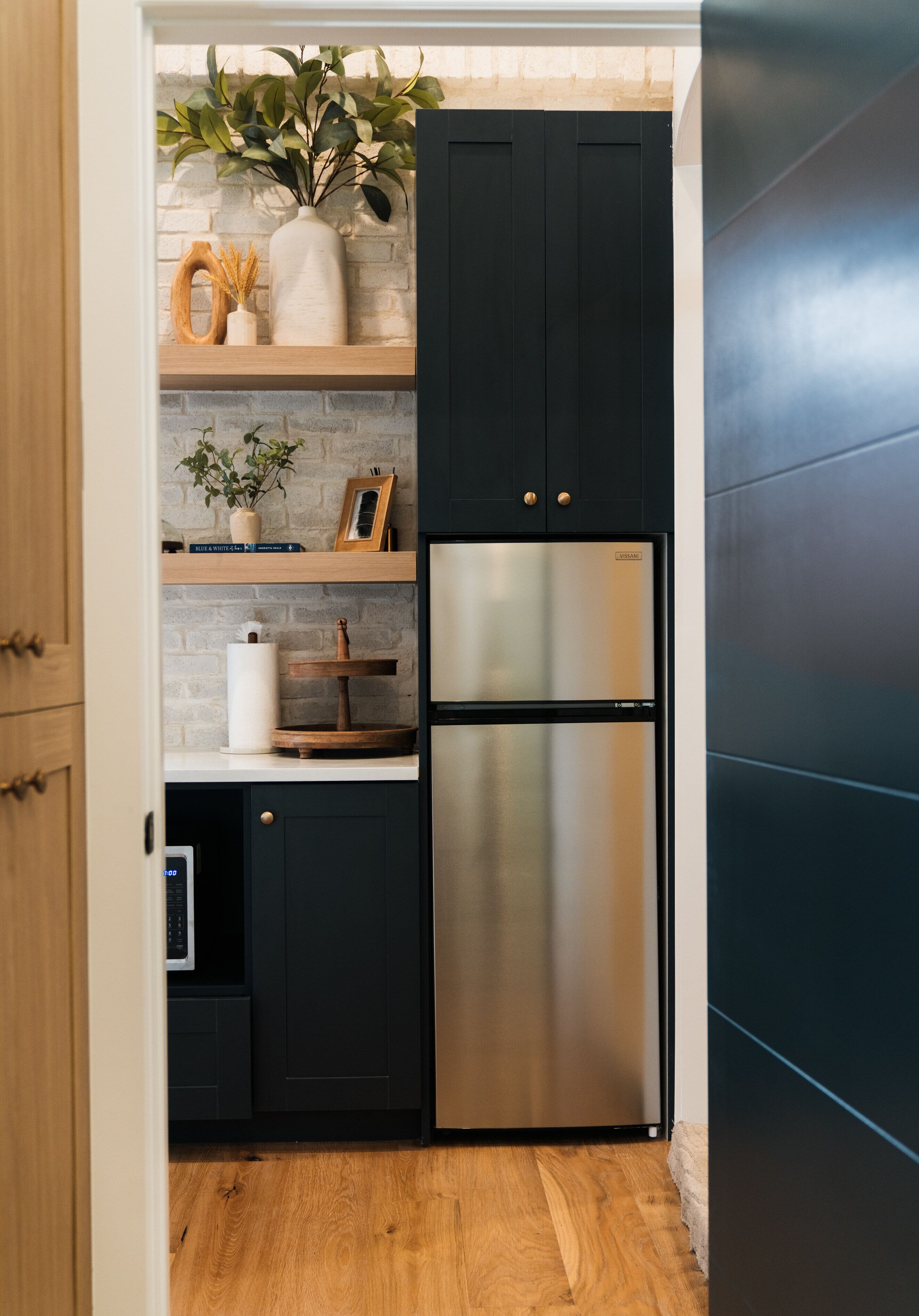 A compact kitchen with dark cabinetry, a stainless steel refrigerator, and wooden shelves.