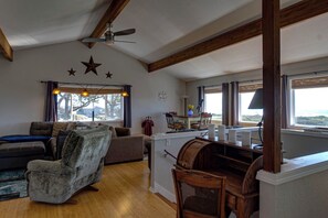 Uma sala de estar acolhedora com um sofá, poltrona e mesa de centro de madeira. Há um ventilador de teto, uma janela com cortinas e uma vista para o exterior.
