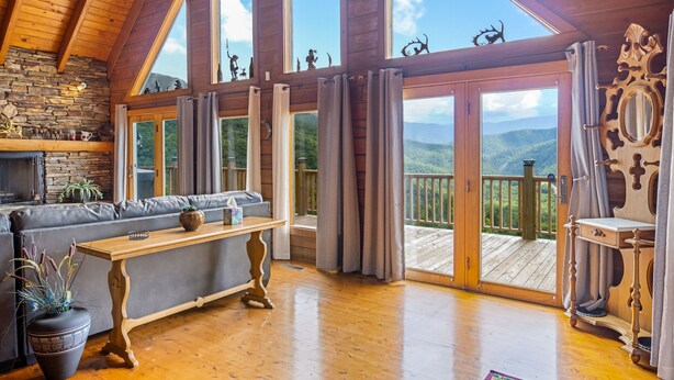 Ein geräumiges Zimmer mit Holzboden, einem Kamin aus Stein, einem grauen Sofa, einem Holztisch und großen Fenstern mit Blick auf die Berge.