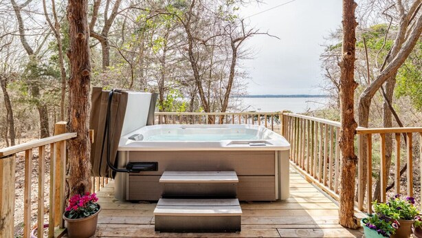 Een hottub op een houten terras, met treden die er naartoe leiden, omringd door bomen en met uitzicht op een wateroppervlak.