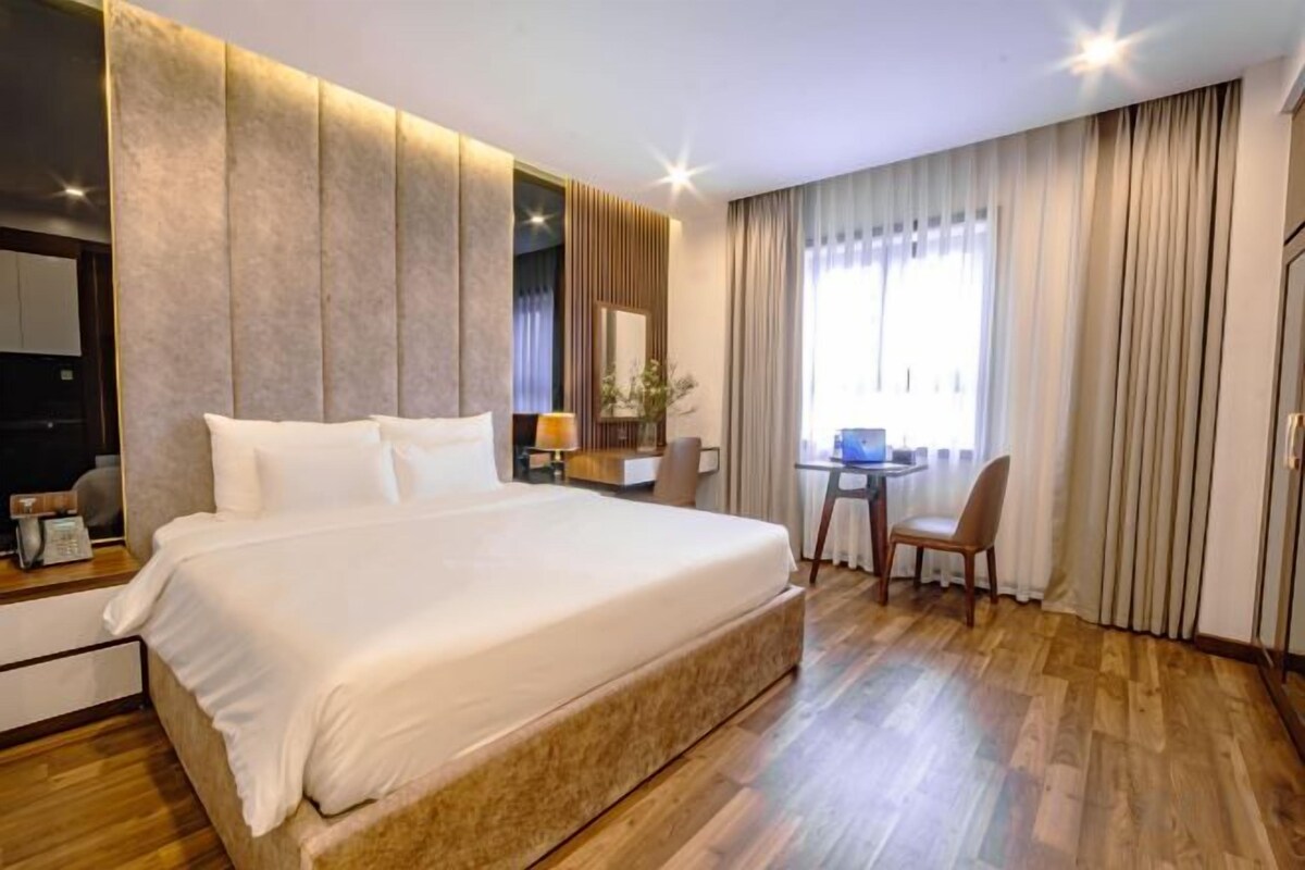 Un grand lit avec une tête de lit en bois, une table de chevet avec une lampe et une chaise dans une chambre d’hôtel dotée d’un plancher en bois et d’une grande fenêtre avec des rideaux.