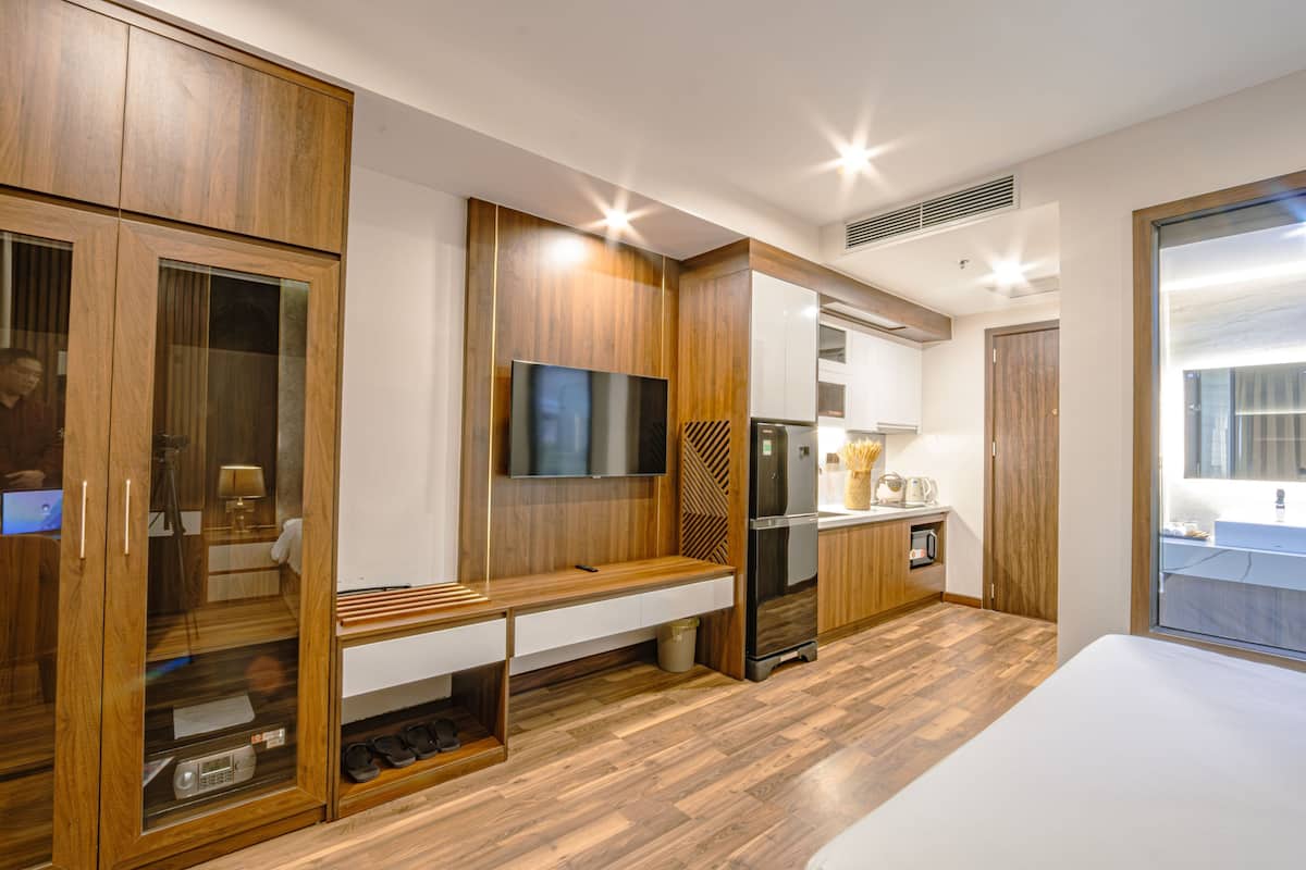 A modern hotel room with wooden cabinetry, a flat-screen TV, a built-in bench, a refrigerator, and a large window offering a view.