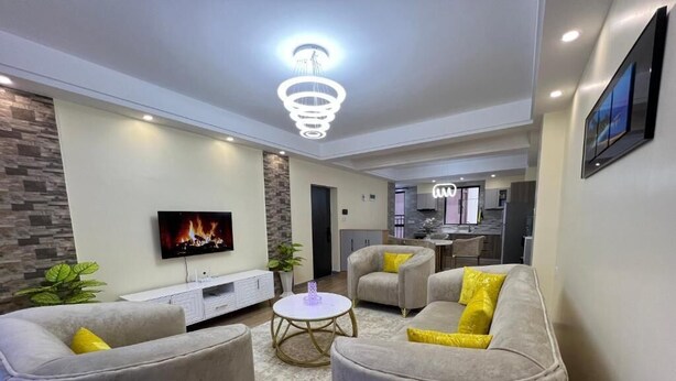Una sala de estar moderna con un televisor de pantalla plana, sofás grises con cojines amarillos, una mesa de centro redonda y una chimenea.