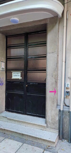 En byggnadsentré med dubbeldörrar, en skylt med texten ”AEF ALEXOMATTE DIACHMITIKA” och en telefonkiosk till höger.